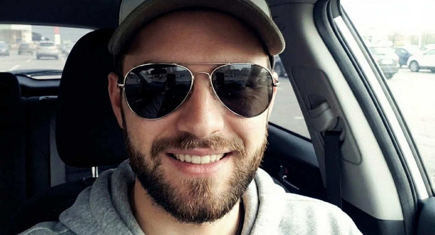 A smiling man wears sunglasses and a cap in a car with visible parking lot background.