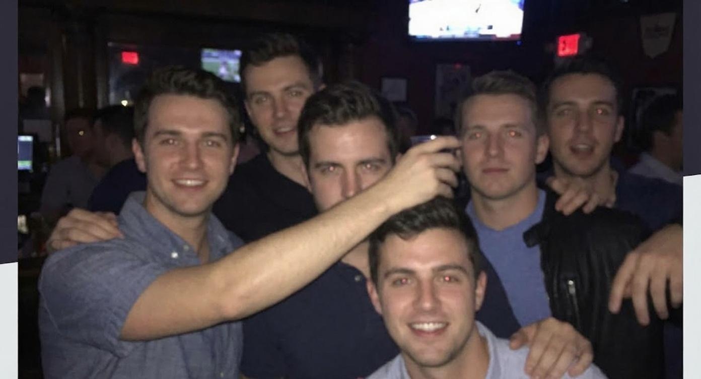 Six young men are clustered together smiling in a dimly lit crowded bar.