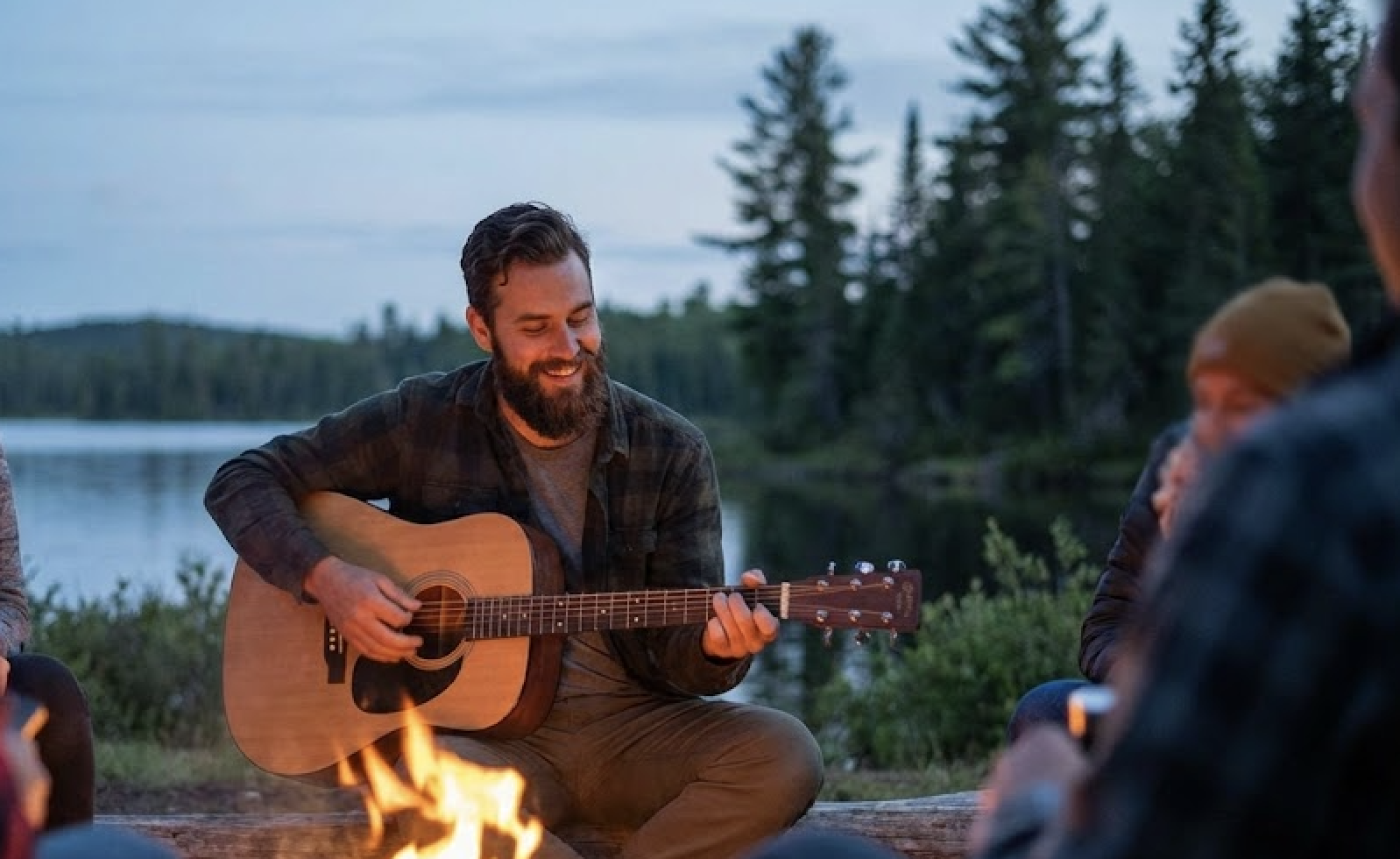 A bearded man smiles while playing guitar by a campfire near a lake with friends.