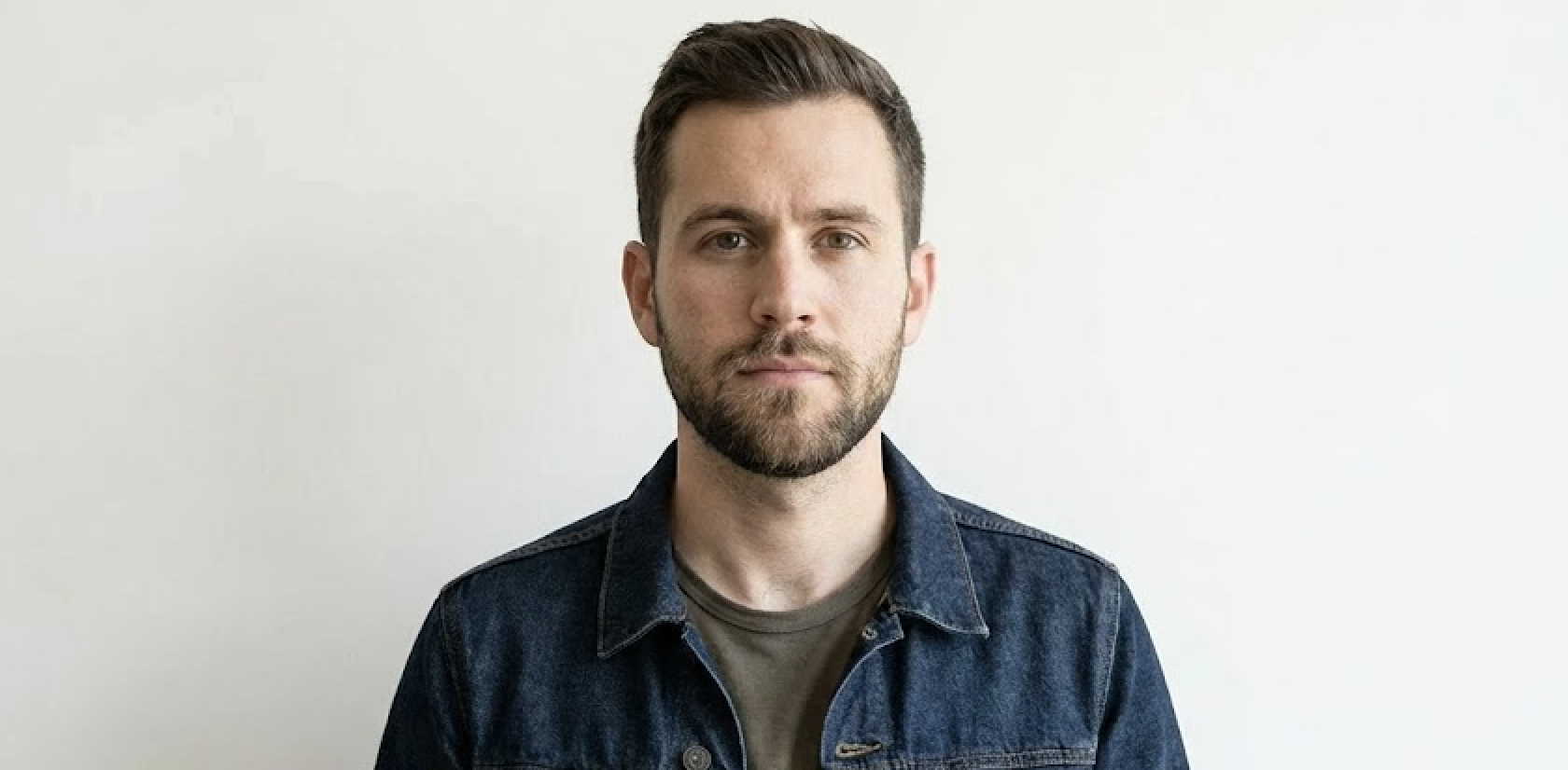 A medium shot shows a Caucasian man against a white wall wearing a dark jacket.