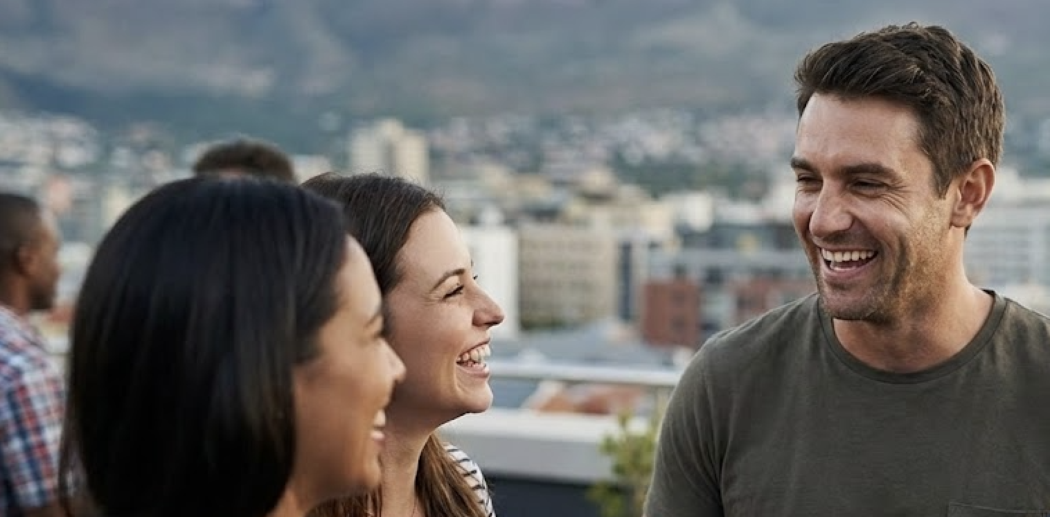 Three people are shown laughing on a rooftop with a city skyline in the background.