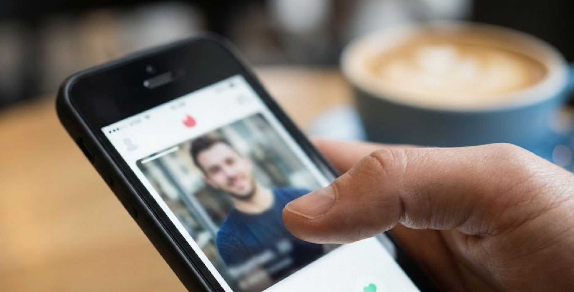 A hand is shown using a dating app on a smartphone with a cup containing a hot beverage.