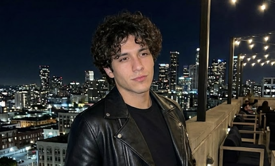 A young man in a leather jacket is posing on a rooftop with a nighttime city skyline behind him.