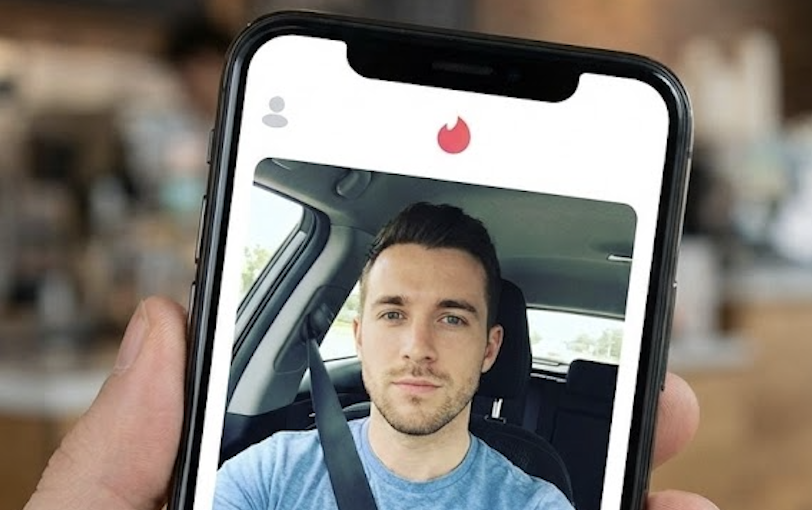 A hand holds a phone displaying a man's photo on the Tinder dating app.