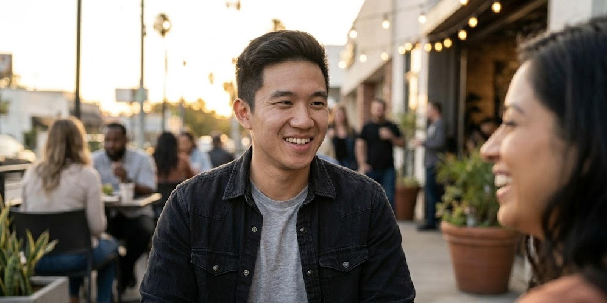 A young man and woman are smiling and talking while sitting at an outdoor cafe.