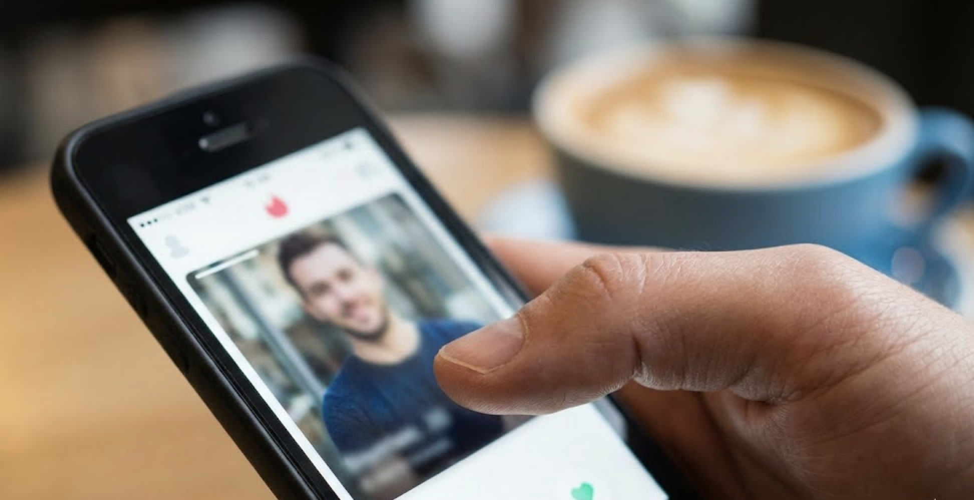 A hand holds a smartphone displaying a dating profile with a man's photo, with coffee blurred behind.