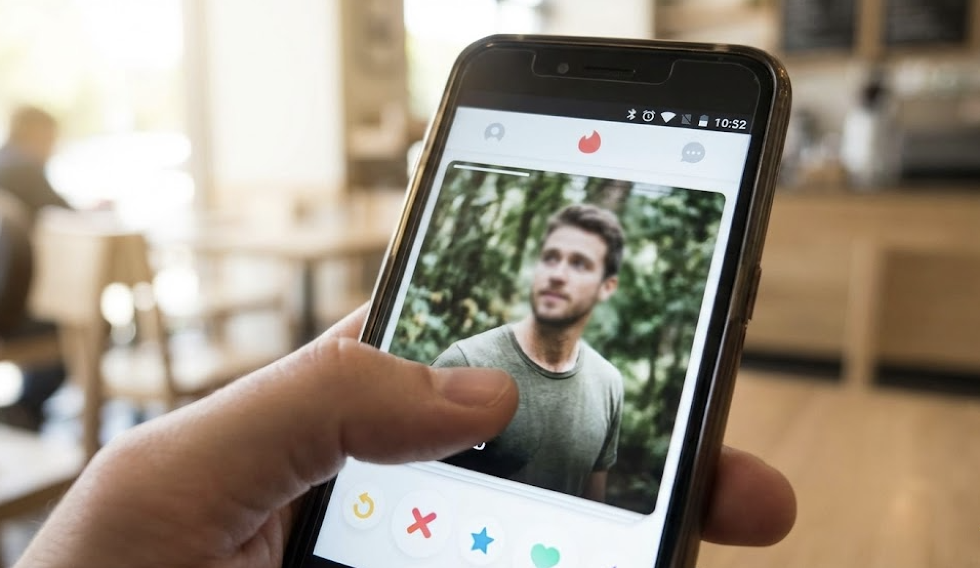 A hand holds a phone showing a male profile on the Tinder dating app.