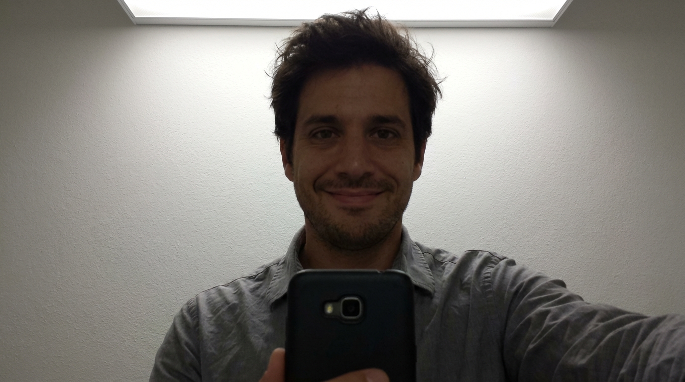 A man with a slight smile holds up a black smartphone indoors against a textured, white wall.