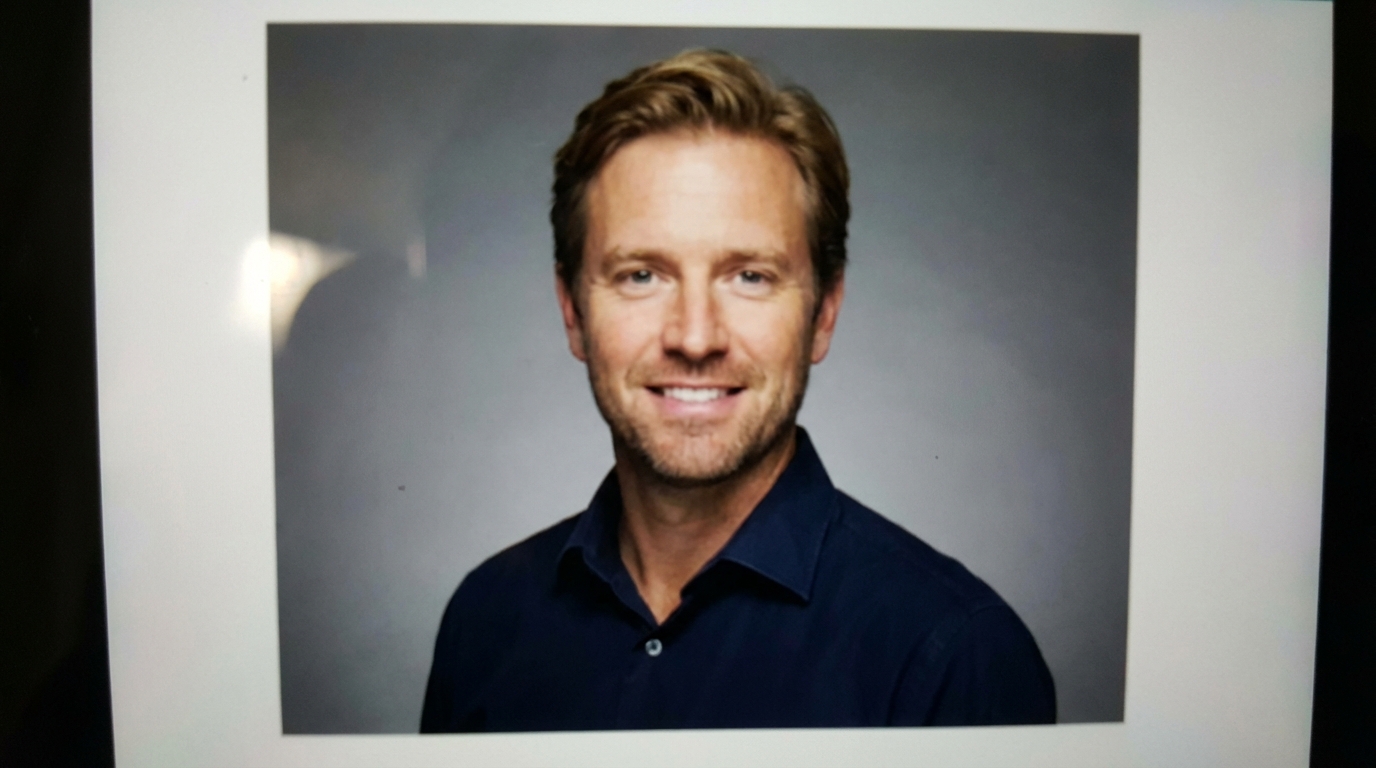 The photo portrays a smiling blond man wearing a blue buttoned shirt against a gray background.