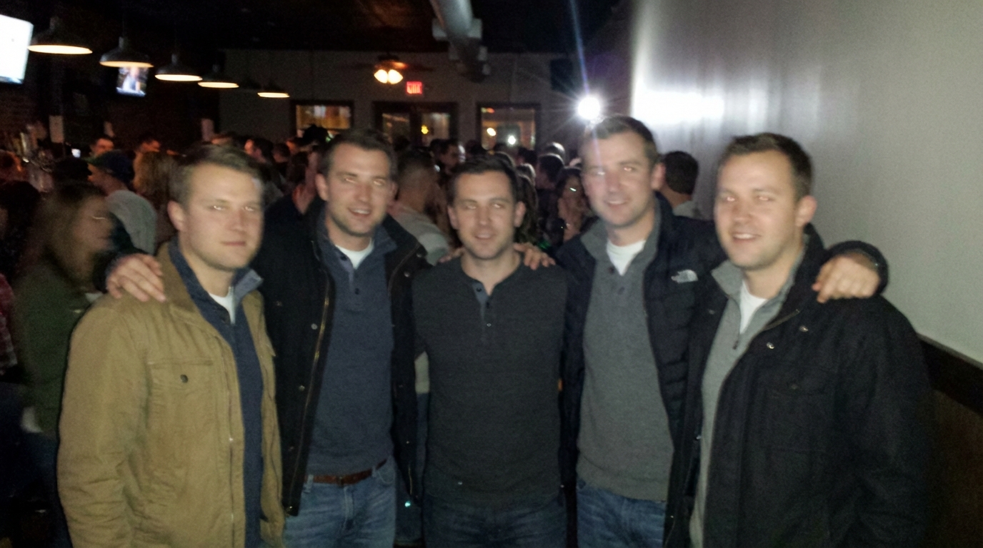 Five young men are posed at a bar, arm in arm, with a busy bar area in the background.