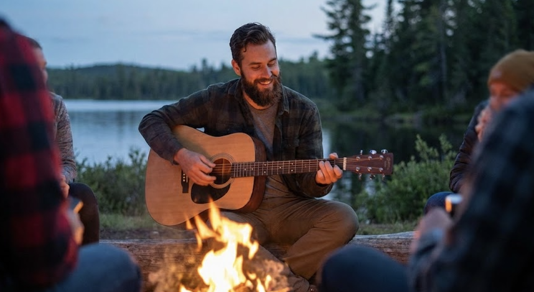 A bearded man plays the guitar for friends around a campfire with a lakeside forest in the background.