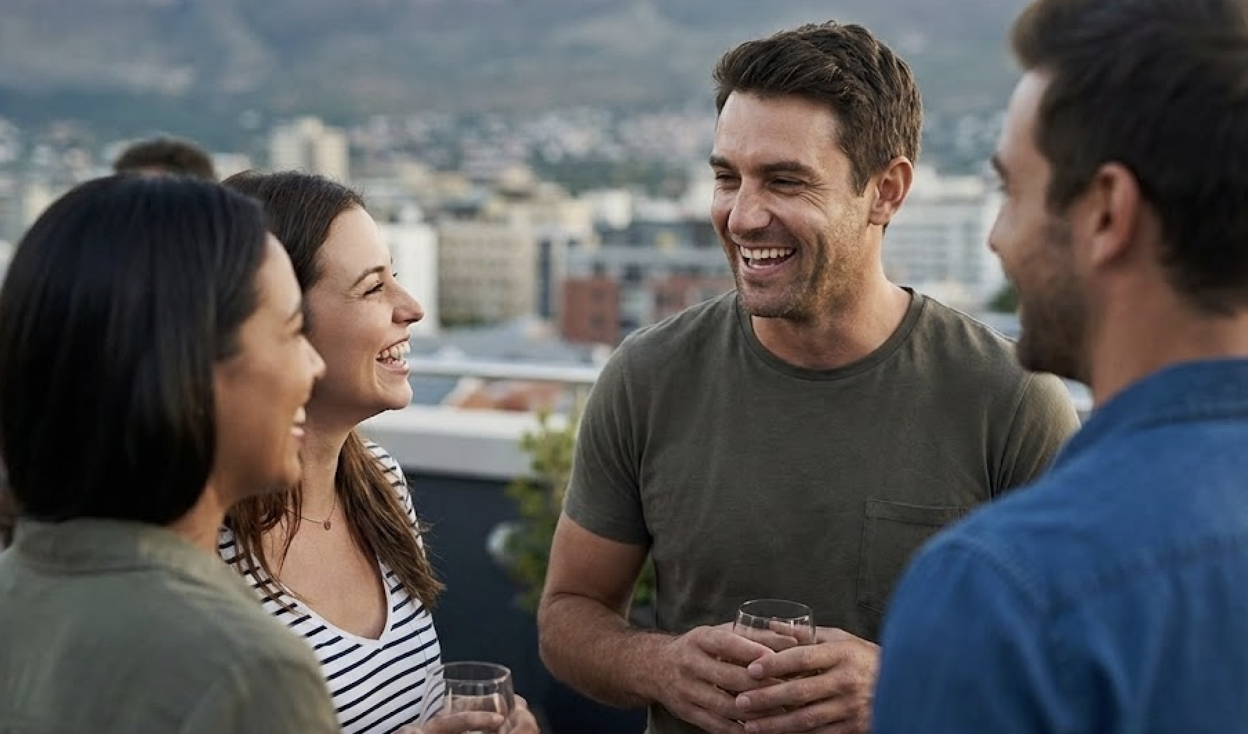 Four adults are laughing and enjoying drinks while standing on a rooftop with a city view.