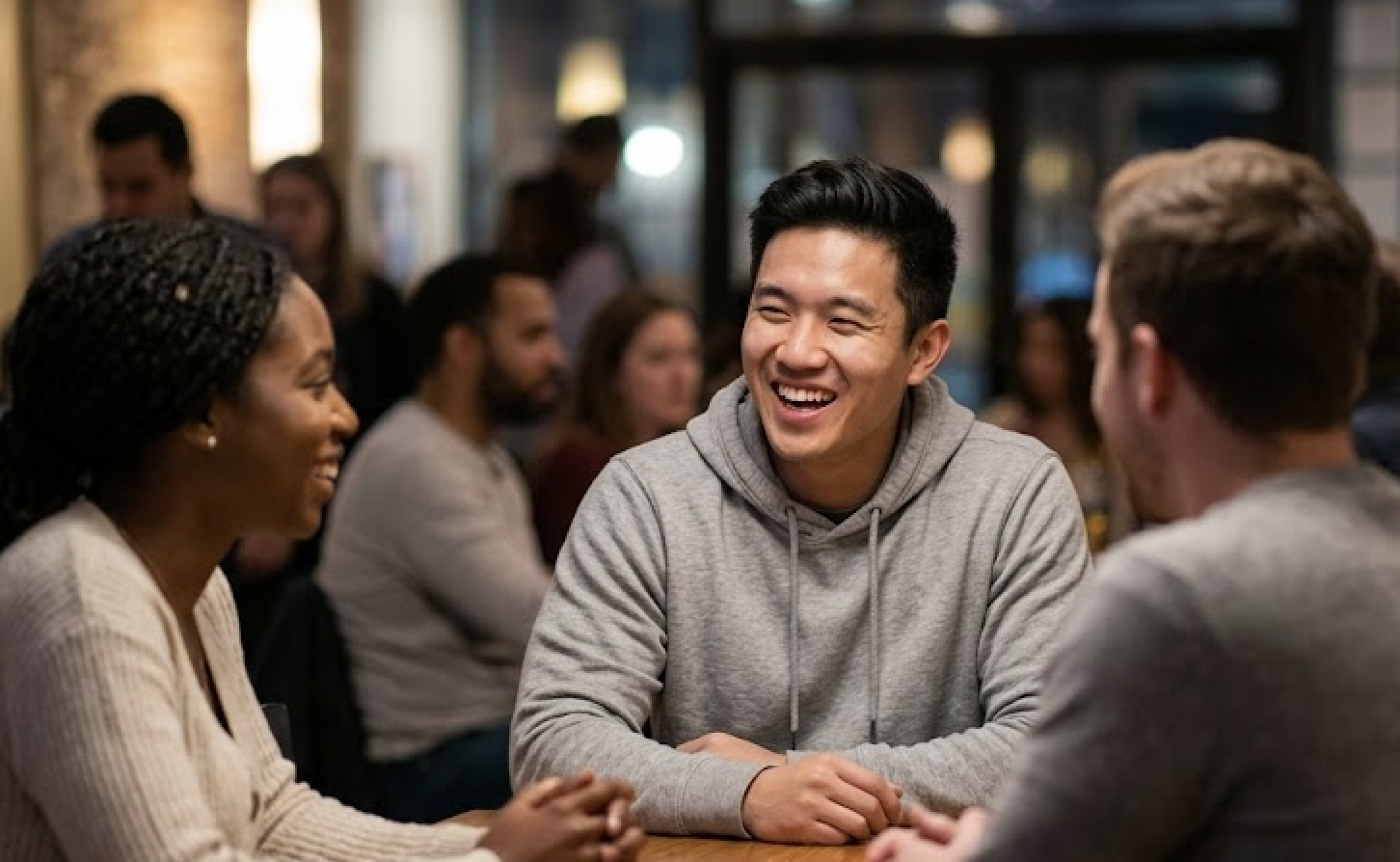 At a wooden table, three smiling people are talking with blurred people in the background.