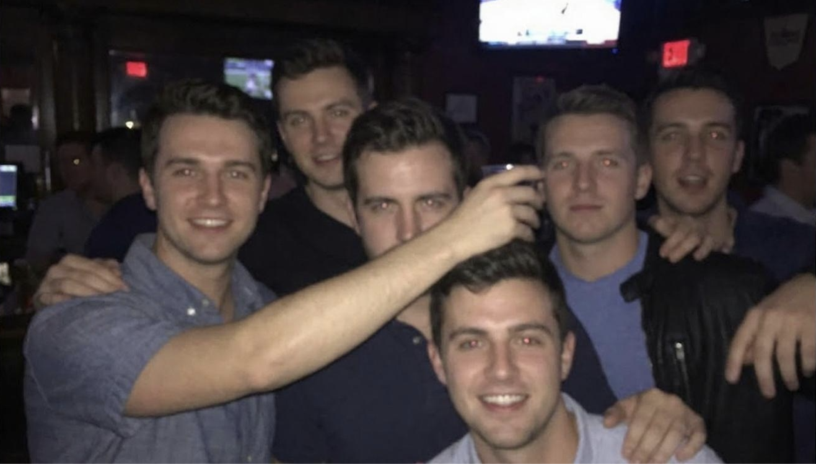 A group of six young men pose together at a dimly lit bar.