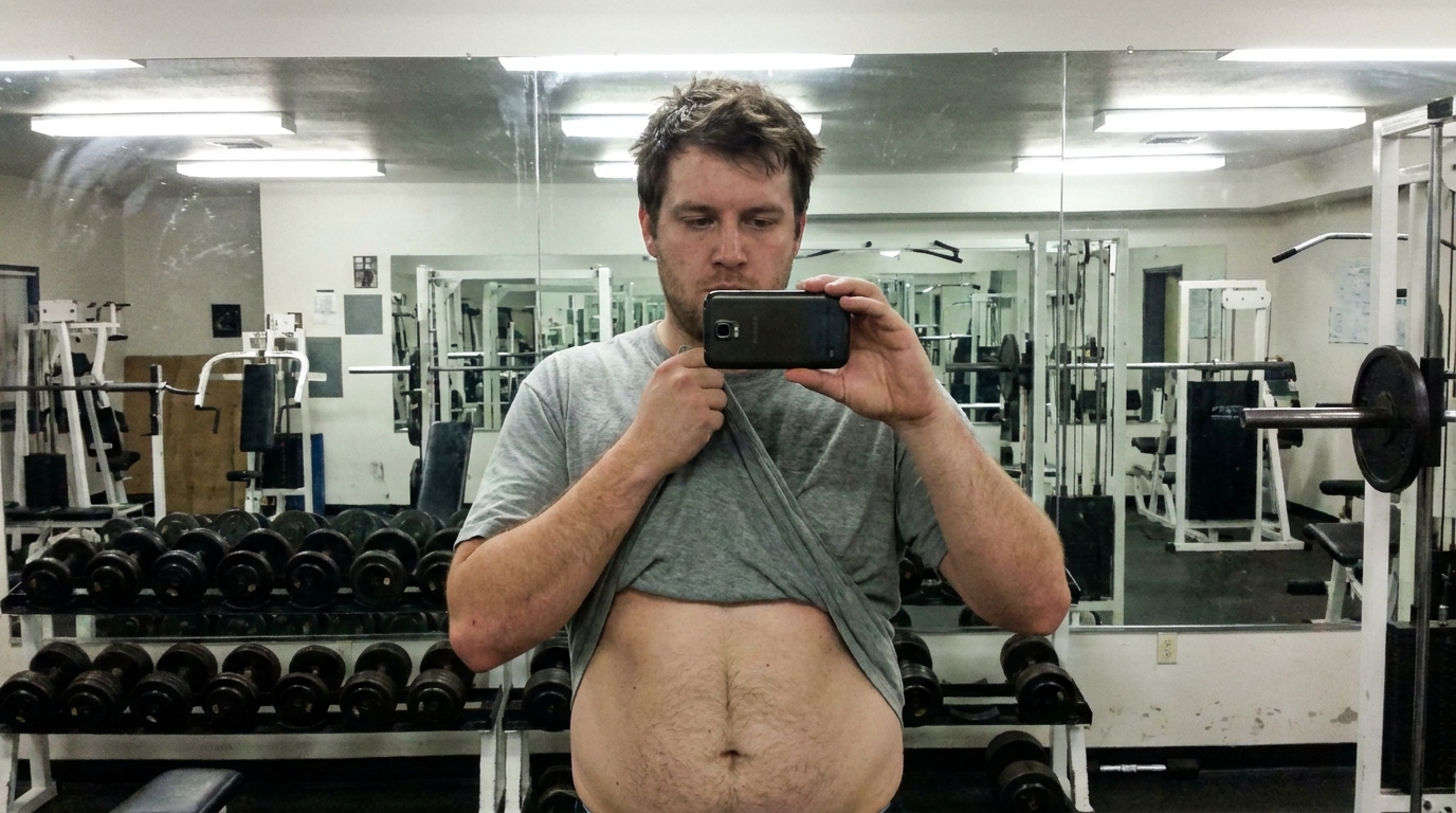 A man holds up his shirt to reveal his belly in a gym locker room.