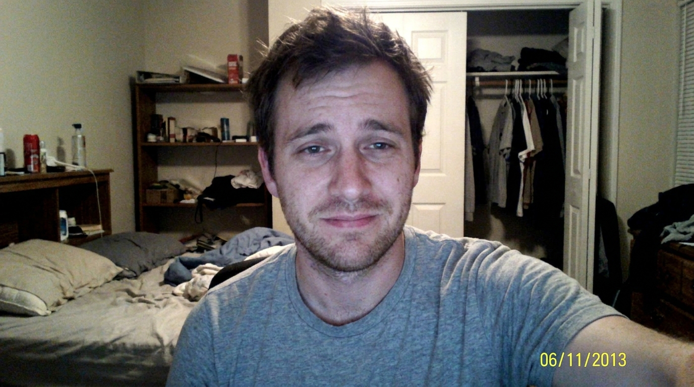 A man with scruffy hair and stubble smiles faintly at the camera in a messy bedroom.
