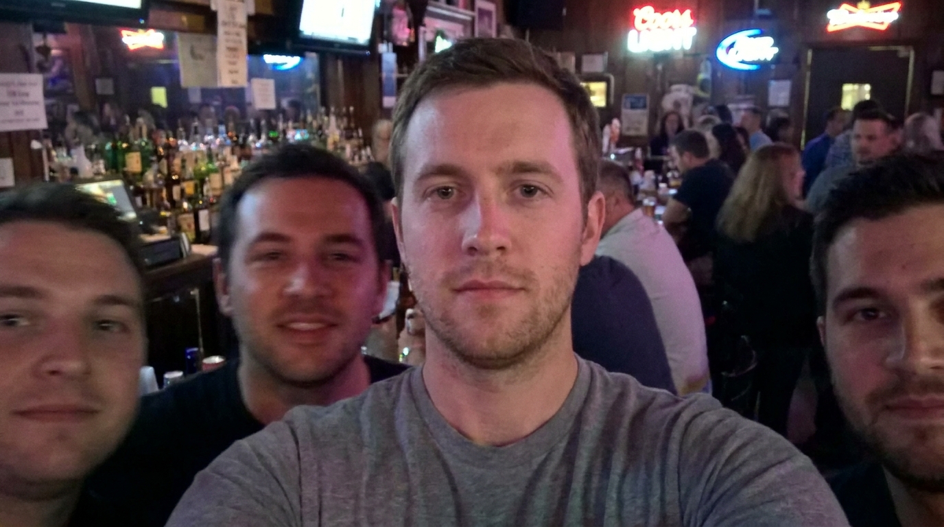Four men take a selfie in a dimly lit bar with neon signs and bottles.