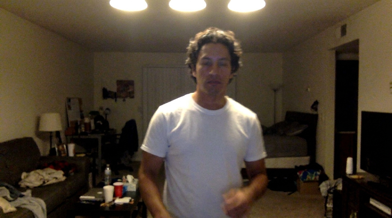 A man with curly hair and a white t-shirt stands in a dimly lit apartment.