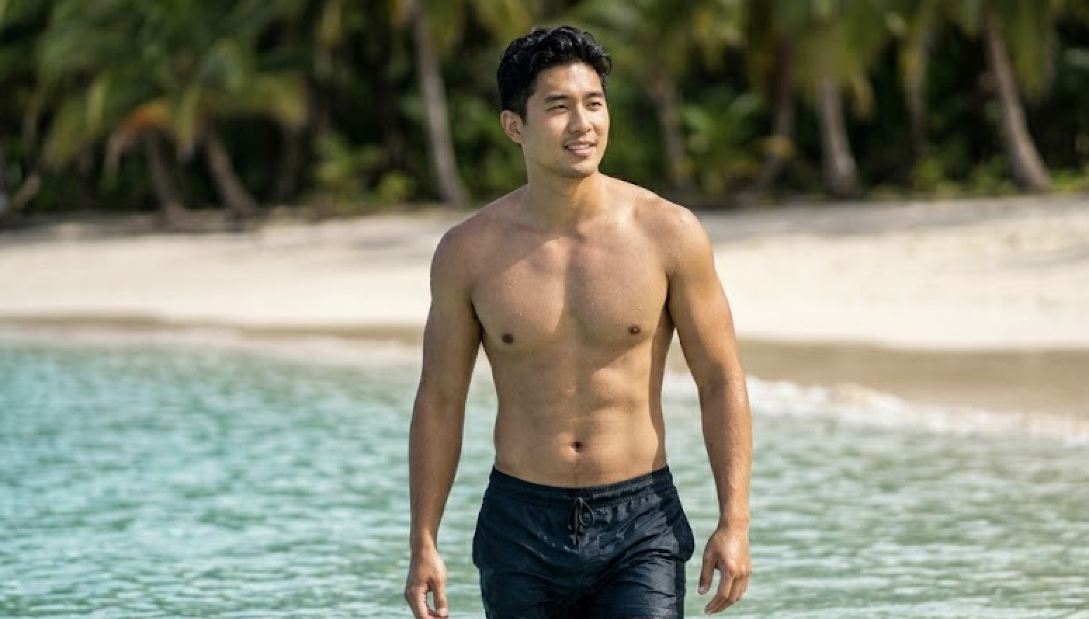 A shirtless Asian man in swim trunks stands in shallow ocean water.
