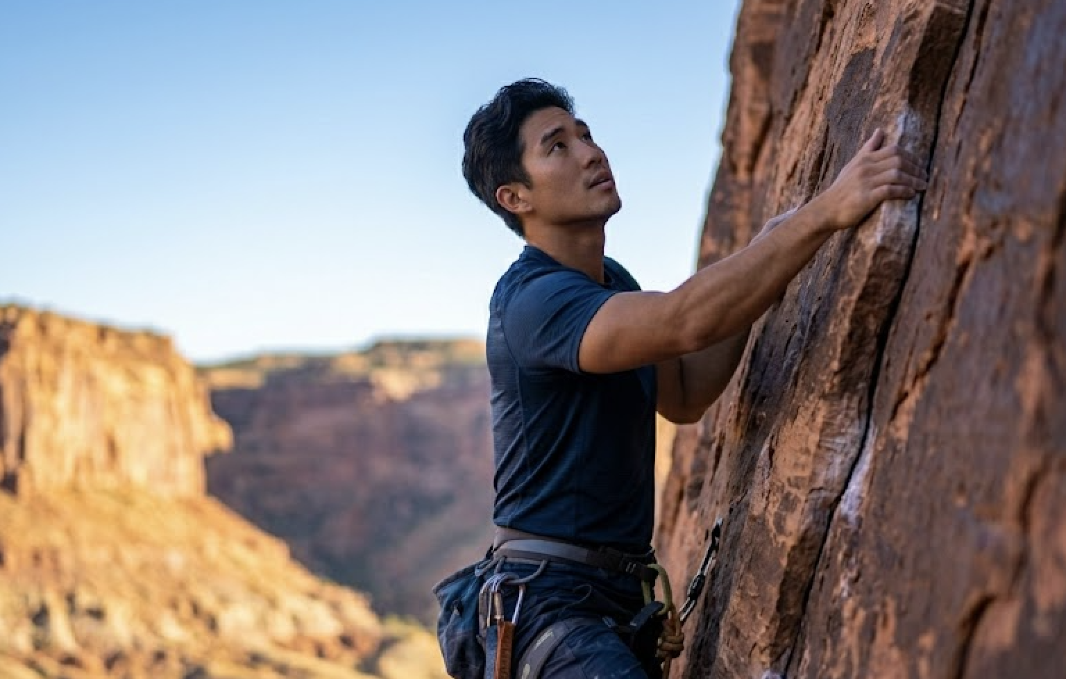 An Asian man in a blue shirt is rock climbing on a brown rock face.