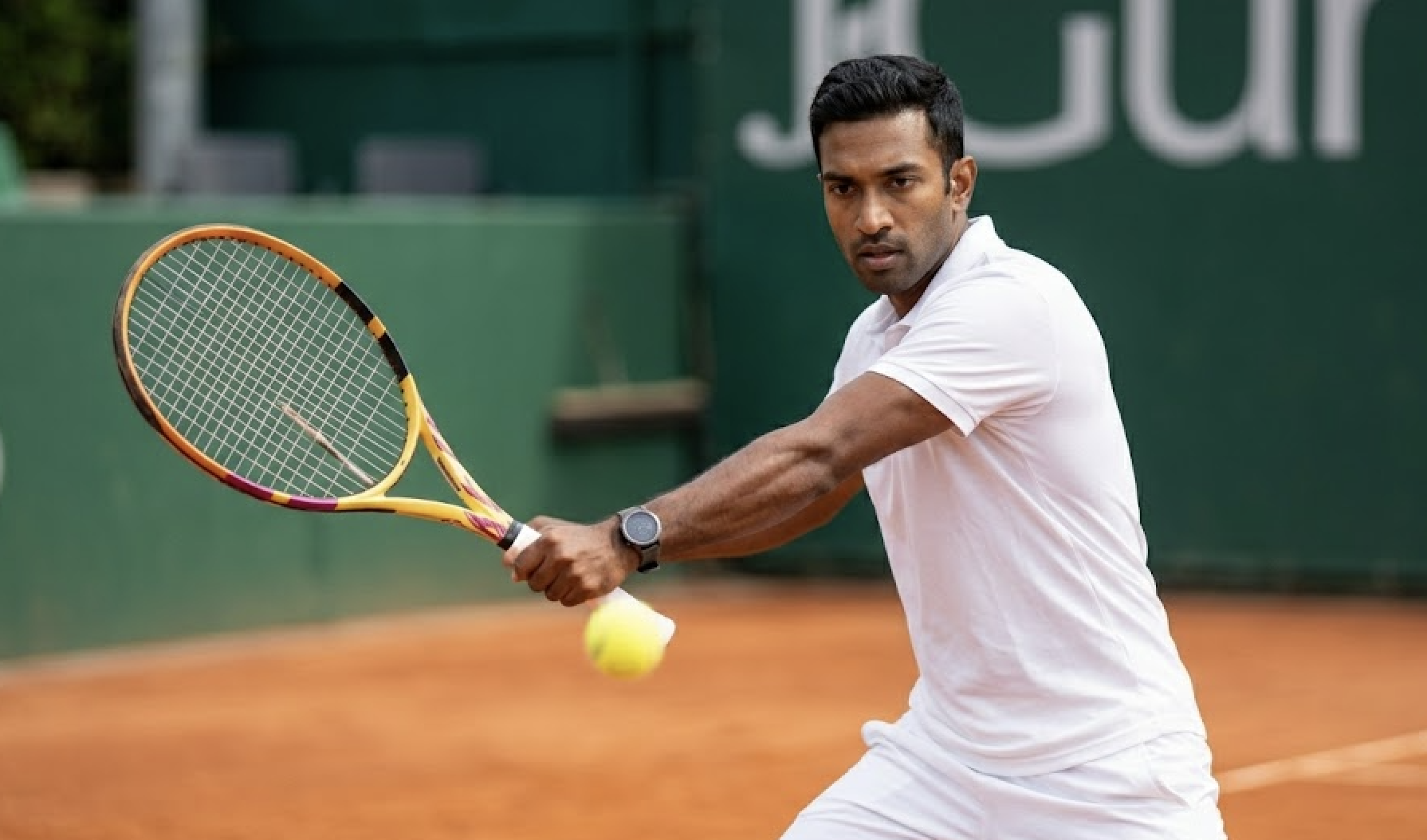 A man in white sportswear plays tennis on a clay court.