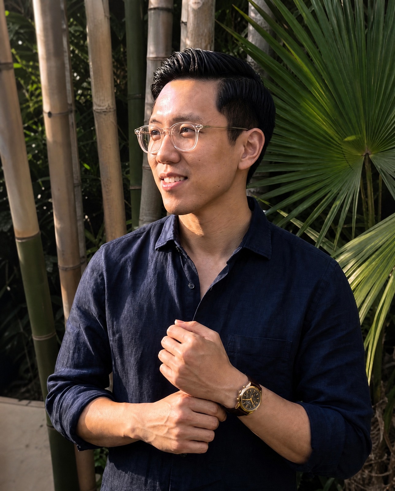 Smiling man with glasses in blue shirt adjusts watch, green plants behind his back.